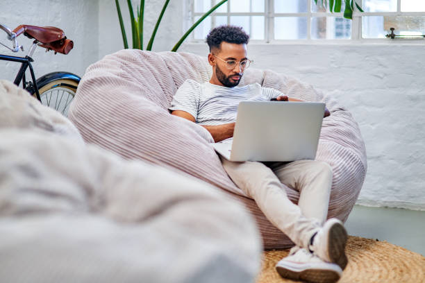 Always modifying my online profile Full length shot of a handsome young businessman sitting on a beanbag alone in his office and using his laptop bean bag stock pictures, royalty-free photos & images