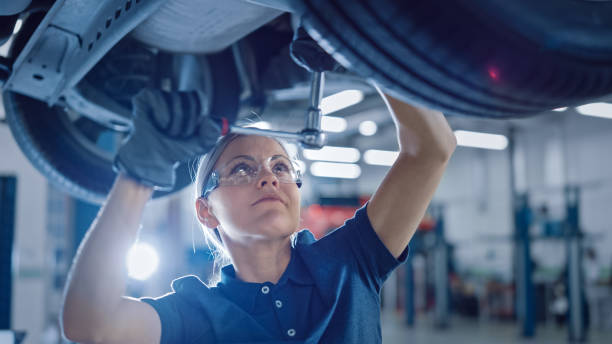 fotografía de un mecánico femenino que trabaja bajo vehículo en un servicio de coche. empoderar a la mujer usando guantes y usar un trinquete debajo del coche. taller limpio moderno. - mechanic fotografías e imágenes de stock