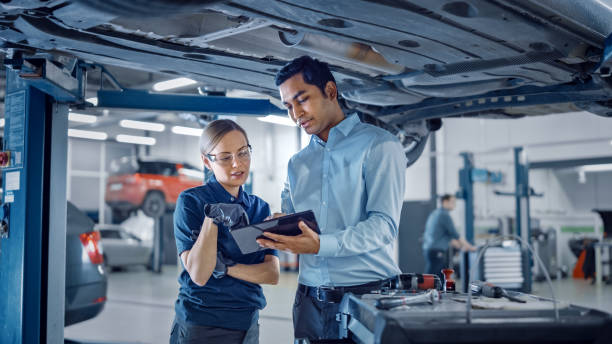 female mechanic talking to a manager under a vehicle in a car service. specialist is showing info on a tablet computer. empowering woman wearing gloves and safety gloves. modern clean workshop. - car equipment smiling working imagens e fotografias de stock