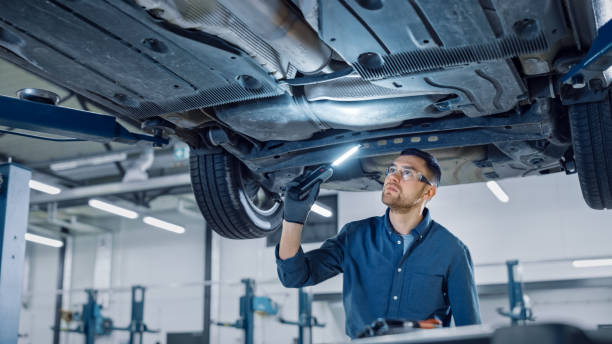 handsome professional car mechanic está investigando el óxido bajo un vehículo en un ascensor en servicio. repairman está utilizando una lámpara led y camina hacia. el especialista está usando gafas de seguridad. taller moderno. - debajo de fotografías e imágenes de stock