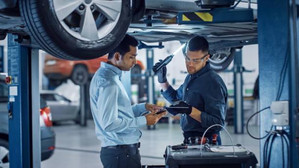 Manager Checks Data on a Tablet Computer and Explains the Breakdown to a Mechanic. Car Service Employees Inspect the Bottom of the Car with a LED Lamp. Modern Clean Workshop.