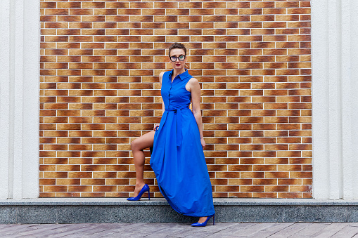 A full-size photo of an attractive woman in a long dress on the high heels on the brick wall background. The woman in glasses poses to a photographer.