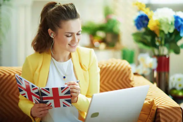 smiling elegant middle age woman in jeans and yellow jacket with laptop taking notes with a pen in a UK flag notebook in the modern house in sunny day.