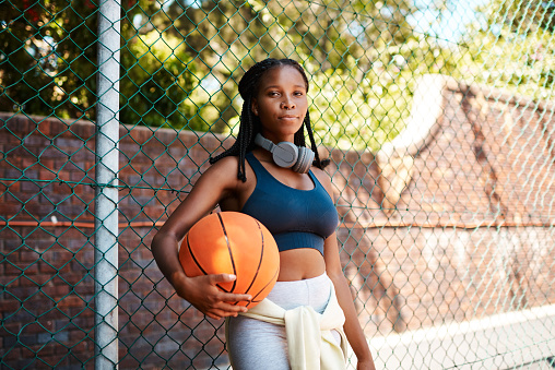 Portrait of a sporty young woman holding a basketball while standing against a fence outdoors