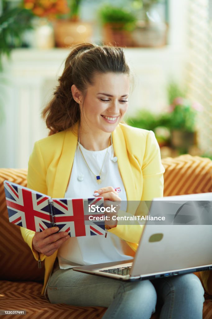 happy learner woman with laptop taking notes in notebook happy elegant learner woman in jeans and yellow jacket with laptop taking notes with a pen in a UK flag notebook in the modern living room in sunny day. England Stock Photo