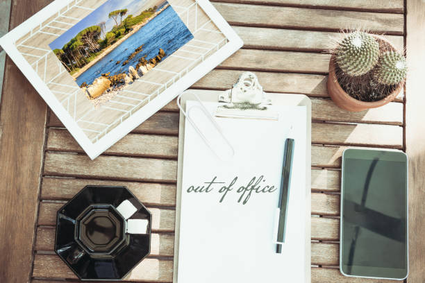 Desk in flat lay with a holiday photo a coffee a smartphone and a cactus the words Out of office written in English on a notepad with his wooden desk pen stock photo