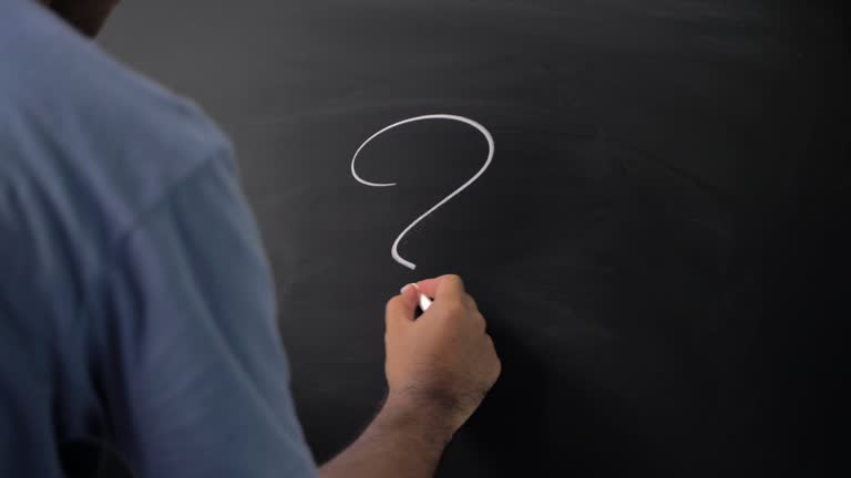 Close up male hand writing question mark with chalk on chalkboard.