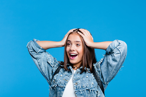 Happy cute teenager wearing oversized denim jacket and white t-shirt standing with raised arms and hands against blue background. Portrait of smiling girl.