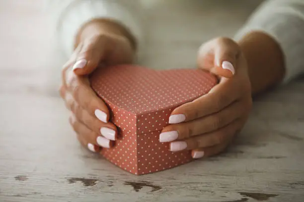 Close up, high angle view of pink heart-shaped gift box placed on a table and held by female hands with nail polish.