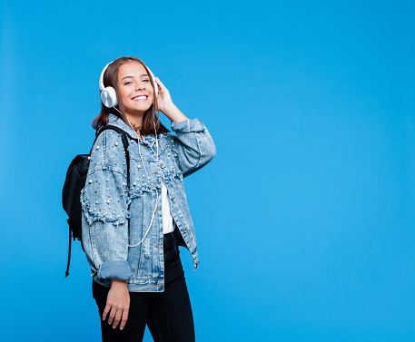 Happy cute teenage girl wearing oversized denim jacket, white t-shirt, black jeans, backpack and headphones standing against blue background. Portrait of smiling teenager.