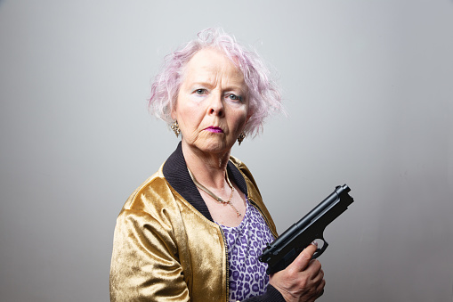 Senior woman, wearing a golden jacket, shirt with leopard pattern, holding gun in her hand, she has pink curly hair
