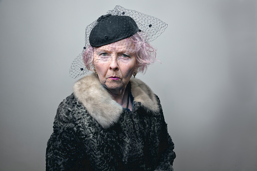 senior woman, wearing fur coat and black hat with veil, with pink hair, standing with a gesture of loneliness, looking with a serious or sad expression