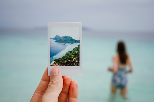 A shot of a hand holding a Polaroid feature the scenery of Sibuan Island in Borneo . Behind the polariod you can see a female walking out to the ocean with her back to camera.