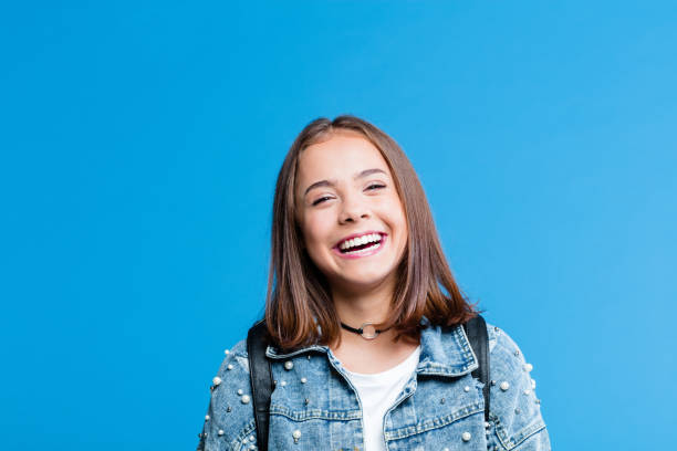 Cheerful pretty teenege girl on blue background Cute female high school student wearing oversized denim jacket and white t-shirt standing against blue background. Portrait of smiling teenager. double denim stock pictures, royalty-free photos & images