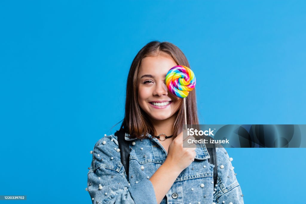 Cheerful teenege girl holding lollipop Cute female high school student wearing oversized denim jacket standing against blue background. Portrait of smiling teenager holding colorful lollipop in front of her eye. Lollipop Stock Photo