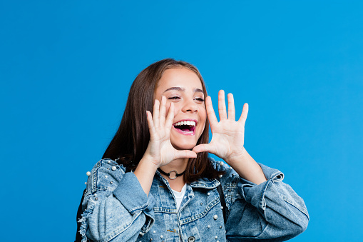 Cute female high school student wearing oversized denim jacket and white t-shirt standing against blue background. Portrait of teenager shouting with raised hands.