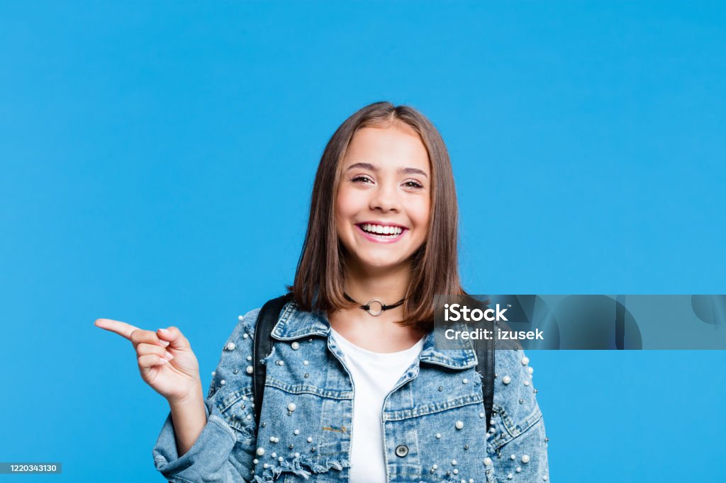 Happy teenge girl pointing with finger at copy space Cute female high school student wearing oversized denim jacket and white t-shirt standing against blue background. Portrait of smiling teenager pointing with index finger at copy space. Teenager Stock Photo