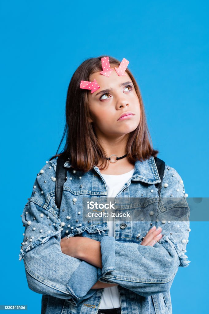 Pensive female hight school student with adhesive note on forehead Teenage girl wearing oversized denim jacket and white t-shirt standing against blue background. Portrait of pensive teenager with with adhesive note on forehead. Choosing Stock Photo