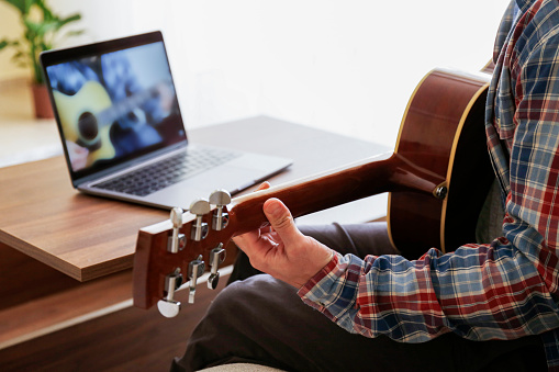Music college hipster student in checkered plaid shirt practicing acoustic guitar exercise, reading notes from laptop computer. Man taking an online musical courses at home during quarantine. Background, close up