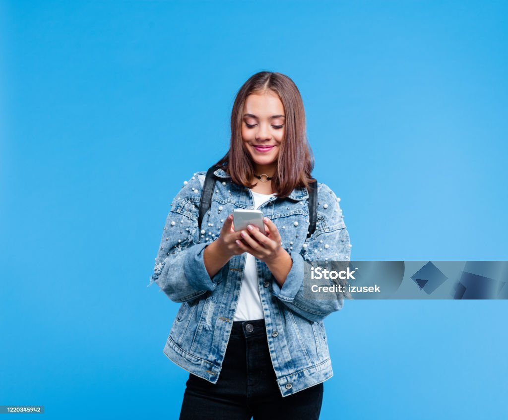 Portrait of female high school student using smart phone Happy teenage girl wearing oversized denim jacket, white t-shirt and black jeans standing against blue background. Portrait of smiling teenager texting on mobile phone. Teenager Stock Photo