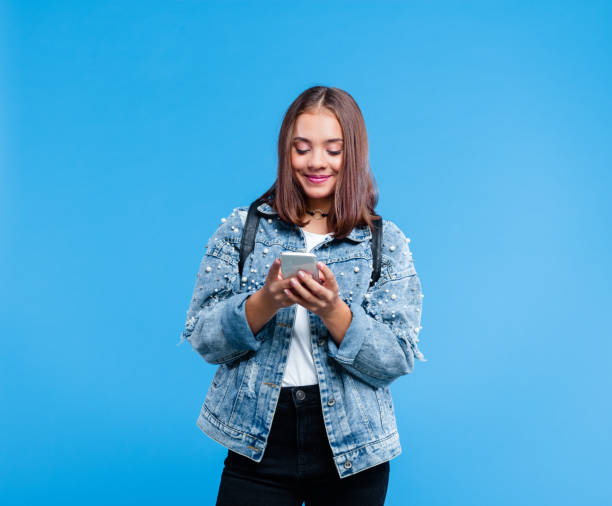 Portrait of female high school student using smart phone Happy teenage girl wearing oversized denim jacket, white t-shirt and black jeans standing against blue background. Portrait of smiling teenager texting on mobile phone. double denim stock pictures, royalty-free photos & images