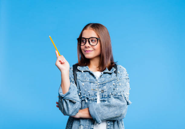 Portrait of petty teenage girl holding pensil Happy teenager wearing oversized denim jacket, white t-shirt and nerdy black eyeglasses pointing with pencil at copy space. Smiling female high school student standing against blue background. double denim stock pictures, royalty-free photos & images