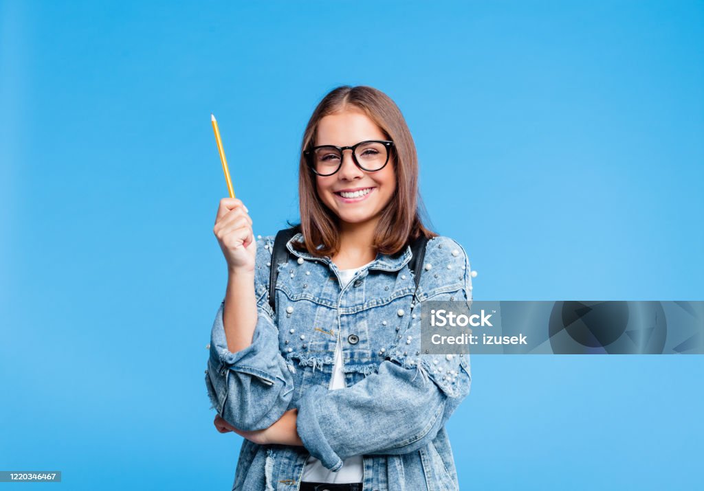 Portrait of petty teenage girl poinitng with pencile Happy teenager wearing oversized denim jacket, white t-shirt and black nerdy eyeglasses pointing with pencil at copy space. Smiling female high school student standing against blue background. Teenager Stock Photo