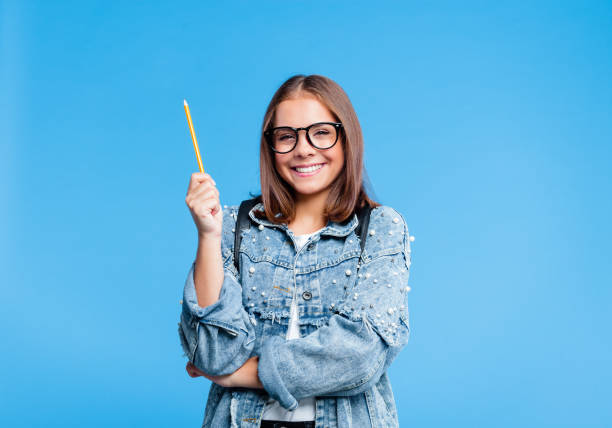 Portrait of petty teenage girl poinitng with pencile Happy teenager wearing oversized denim jacket, white t-shirt and black nerdy eyeglasses pointing with pencil at copy space. Smiling female high school student standing against blue background. double denim stock pictures, royalty-free photos & images