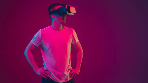 Playing with VR, shoting, driving. Caucasian man's portrait isolated on pink-purple studio background in neon light. Male model with devices. Concept of human emotions, facial expression, sales, ad.