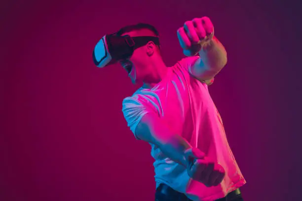 Playing with VR, shoting, driving. Caucasian man's portrait isolated on pink-purple studio background in neon light. Male model with devices. Concept of human emotions, facial expression, sales, ad.