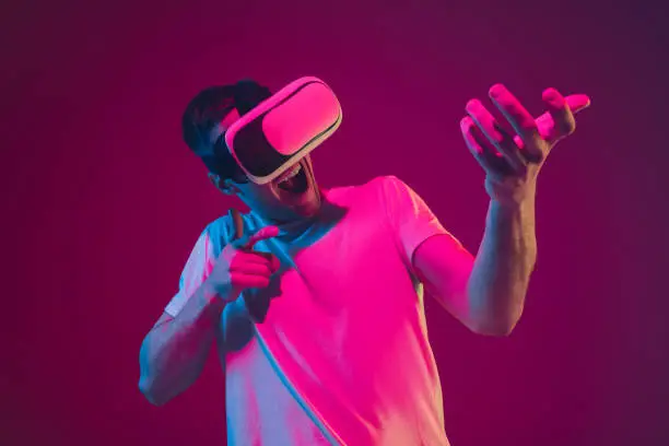 Playing with VR, shoting, driving. Caucasian man's portrait isolated on pink-purple studio background in neon light. Male model with devices. Concept of human emotions, facial expression, sales, ad.