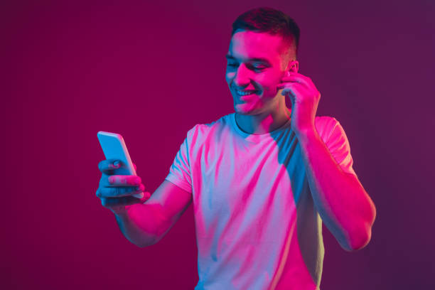 kaukasisches männerporträt isoliert auf rosa-lila studiohintergrund in neonlicht, mit smartphone und kopfhörern - neon fotos stock-fotos und bilder