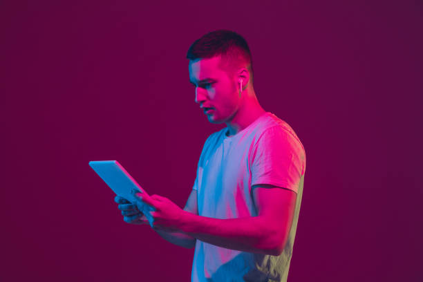 istock Caucasian man's portrait isolated on pink-purple studio background in neon light, using tablet 1220352654