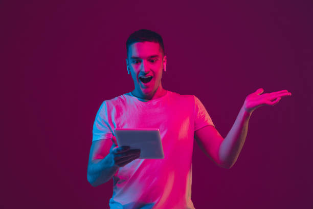 istock Caucasian man's portrait isolated on pink-purple studio background in neon light, using tablet 1220352659