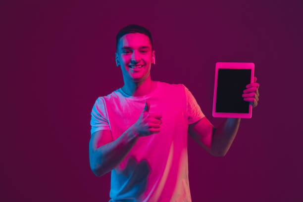 istock Caucasian man's portrait isolated on pink-purple studio background in neon light, using tablet 1220352669