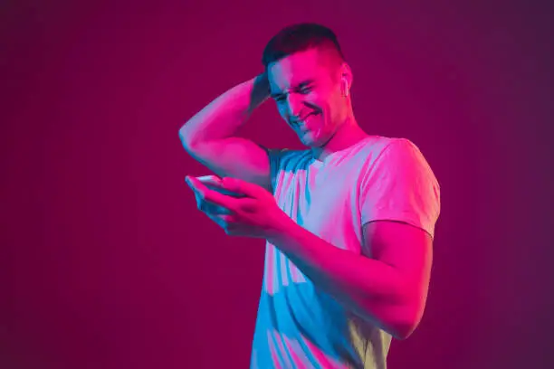 Surfing, betting, watching, selfie. Caucasian man's portrait isolated on pink-purple studio background in neon light. Male model with devices. Concept of human emotions, facial expression, sales, ad.