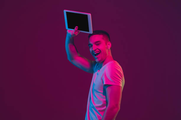 istock Caucasian man's portrait isolated on pink-purple studio background in neon light, using tablet 1220352688