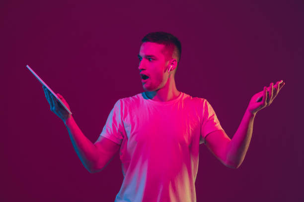 istock Caucasian man's portrait isolated on pink-purple studio background in neon light, using tablet 1220352699