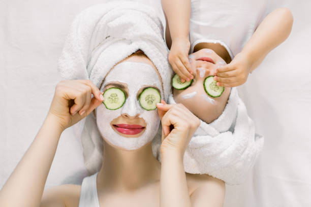 mom and her 2 years old baby girl having fun together, making clay facial mask and cucumber slices on eyes. mother with child doing beauty treatment together. family time, spa and beauty, mothers day - facial mask spa treatment cucumber human face imagens e fotografias de stock