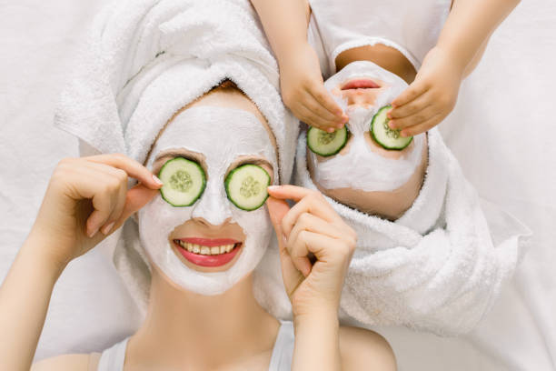 Photo of cheerful mother and little daughter with wrapped hair in white bath towels, white facial mud mask on faces, lying on the bed, and applying pieces of cucumber to their eyes. Family spa Photo of cheerful mother and little daughter with wrapped hair in white bath towels, white facial mud mask on faces, lying on the bed, and applying pieces of cucumber to their eyes. Family spa. women facial mask mud cucumber stock pictures, royalty-free photos & images