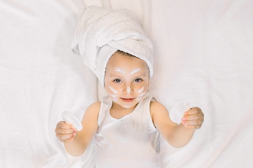 Pretty 2 years old girl child relaxing in the bedroom after shower, lying on the bed, wearing white towel on head and holding in hands two cotton pads. Morning care and baby hygiene.