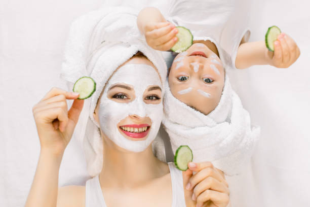 Mother and daughter in white shirts and white towels on their heads in a home bathroom, doing spa procedures with mud mask and fresh cucumber slices. Family time, spa and beauty, mothers day Mother and daughter in white shirts and white towels on their heads in a home bathroom, doing spa procedures with mud mask and fresh cucumber slices. Family time, spa and beauty, mothers day. women facial mask mud cucumber stock pictures, royalty-free photos & images