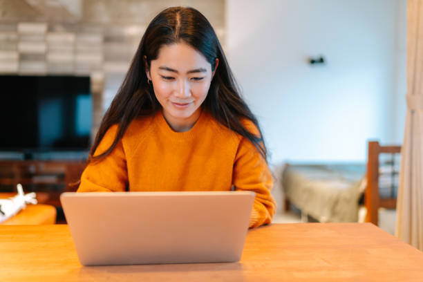 Young stylish woman using laptop at home A young stylish woman is sitting at a table and using a laptop in the living room at home. asian adult student stock pictures, royalty-free photos & images