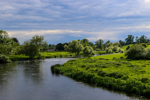 The dark waters of the River Boyne as it meanders through the Irish countryside in summer with lush green foliage along the banks of the river and an overcast cloudy sky above