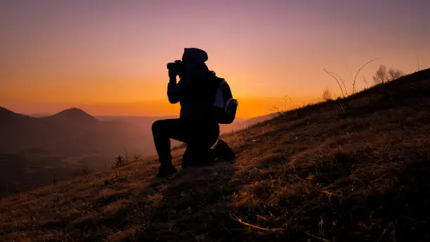 Photo of Very interesting moment. The young photographer managed to climb to the top and achieve his goal. Fantastic sunset and red skies in the background. Active life concept.