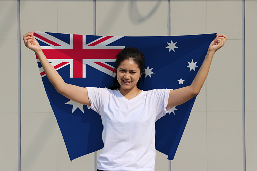 pretty lady is holding Australia flag in her hands and raising to the end of the arm at the back on grey background.
