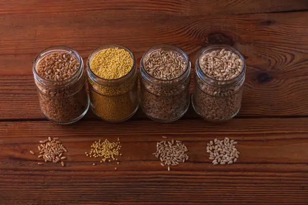 Cereal grains in glass jars on wooden background. Collection of different groats top view barley, oats, millet and wheat. Wholegrain foods with high fiber content flat lay. Healthy diet ingredients.