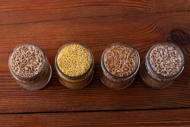 Cereal grains in glass jars on wooden background. Collection of different groats top view barley, oats, millet and wheat. Wholegrain foods with high fiber content flat lay. Healthy diet ingredients.