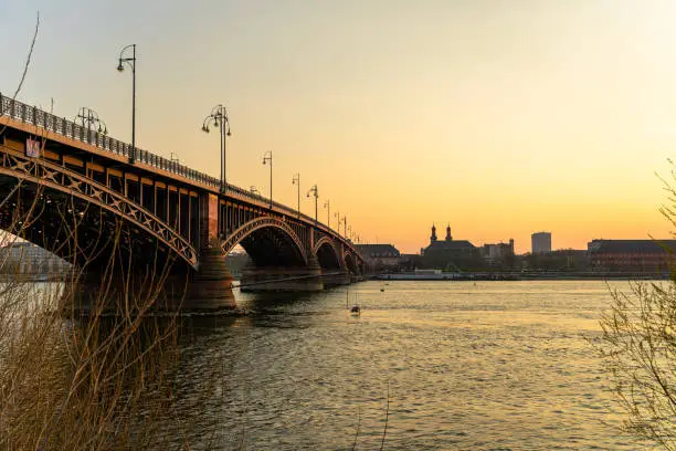 The Theodor-Heuss Bridge spans the golden rhine into the silhouette of the city of Mainz illuminated by the sunset.