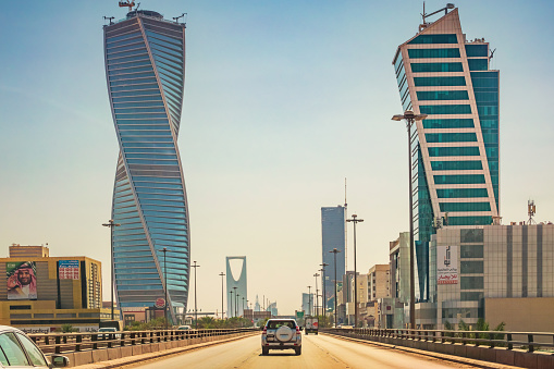 Cars drive below modern skyscrapers on King Fahd Road in Riyadh, Saudi Arabia on a sunny day.